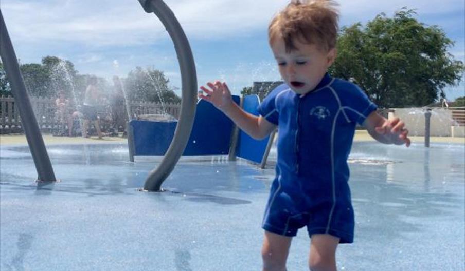 Young child in sunsuit jumping in pool of water with fountains spraying in background