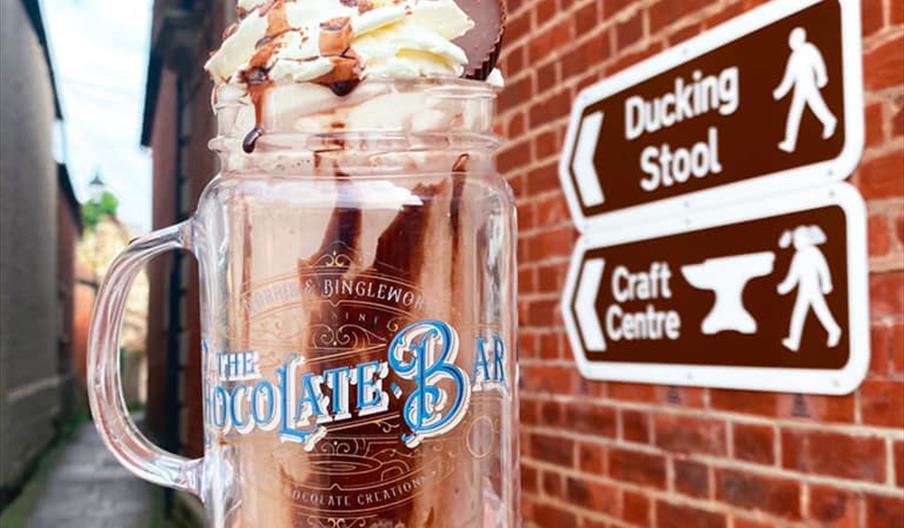 A delicious looking milkshake held aloft next to a brick wall with a brown information sign that points towards the ducking stool.