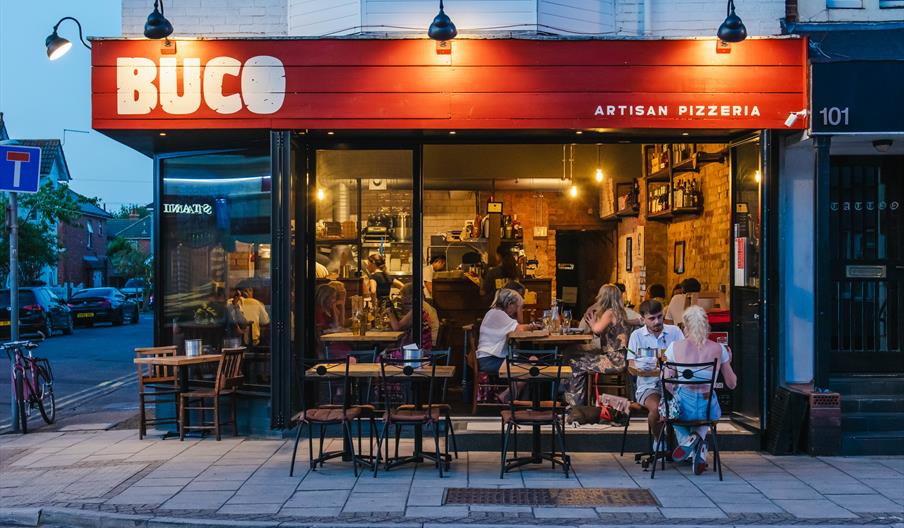 exterior of pizzeria at night, full of people inside and a couple outside