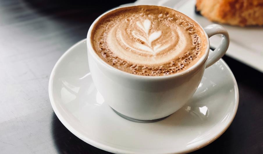 cup of coffee with latte art in a white cup and saucer, with a pastry in the background.