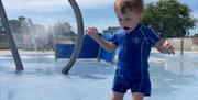 Young child in sunsuit jumping in pool of water with fountains spraying in background