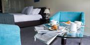 Croissants and coffee on a small table with a newspaper, alongside chairs and a double bed.